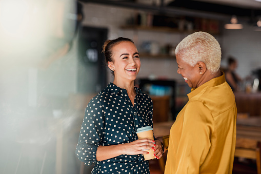 Two women colleagues laughing while standing in a cafe at their workplace. One of the women is holding a take out hot drink cup.