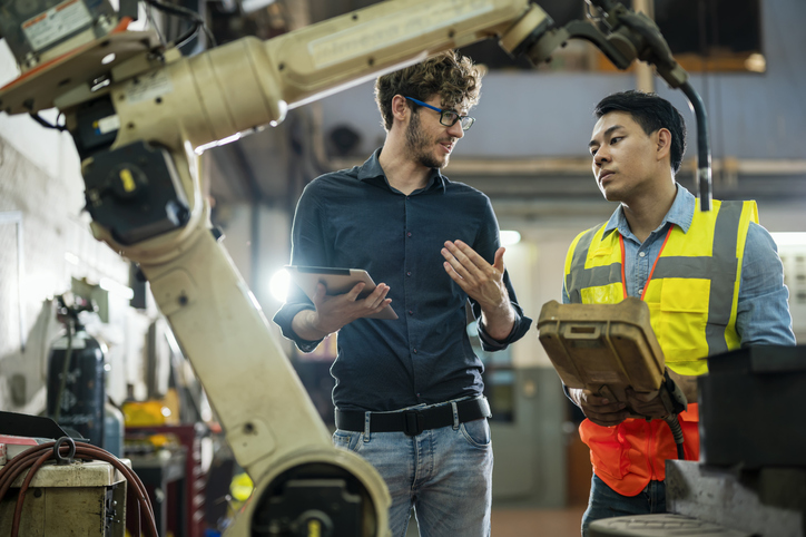 Software engineer explaining to controlling robotic welding process to welder in factory.