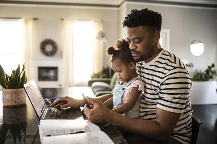 Young Black father looking at computer and phone w young daughter on his lap
